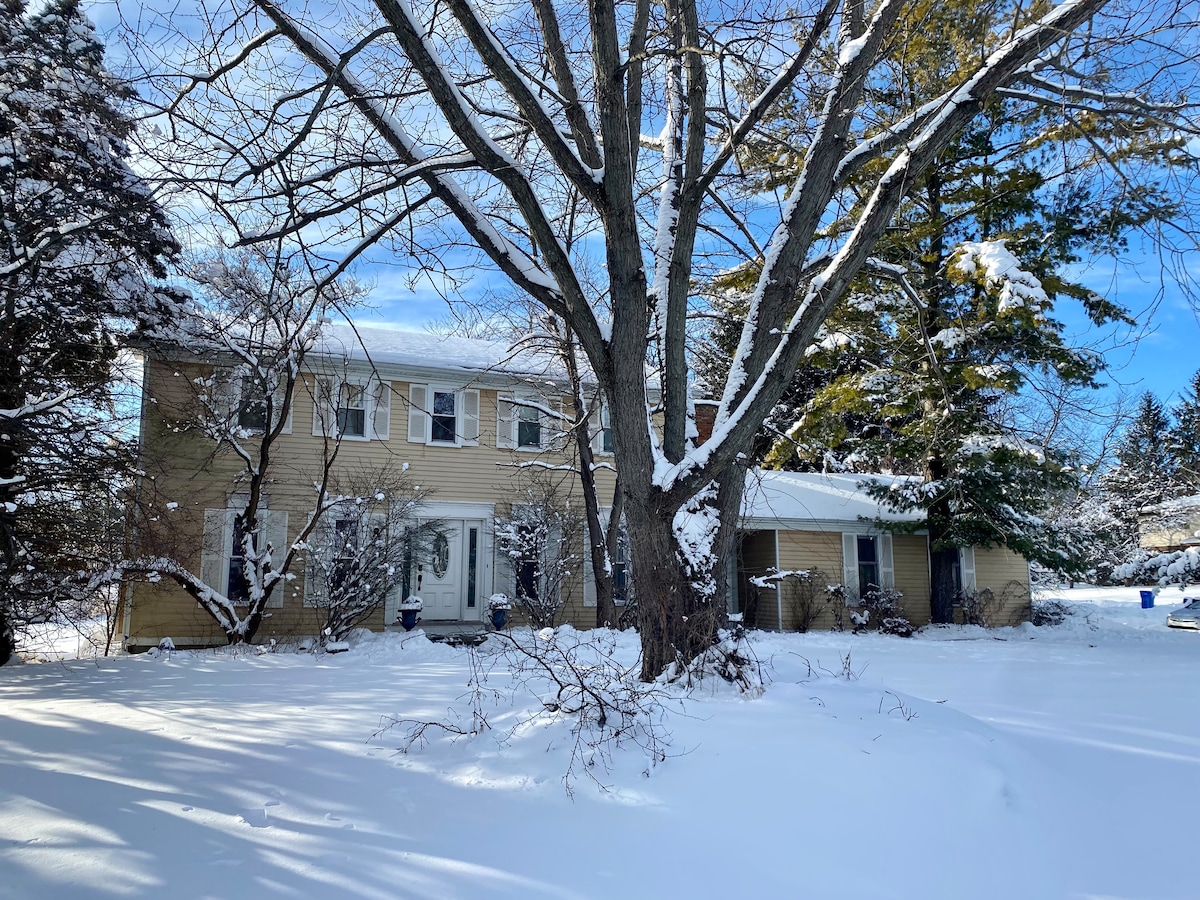 A two-story house is set against a winter landscape, surrounded by a blanket of fresh snow. Leafless trees provide a natural frame, while clear blue skies above add brightness to the scene. The home features a mix of light and dark siding, with a covered porch on the right.
