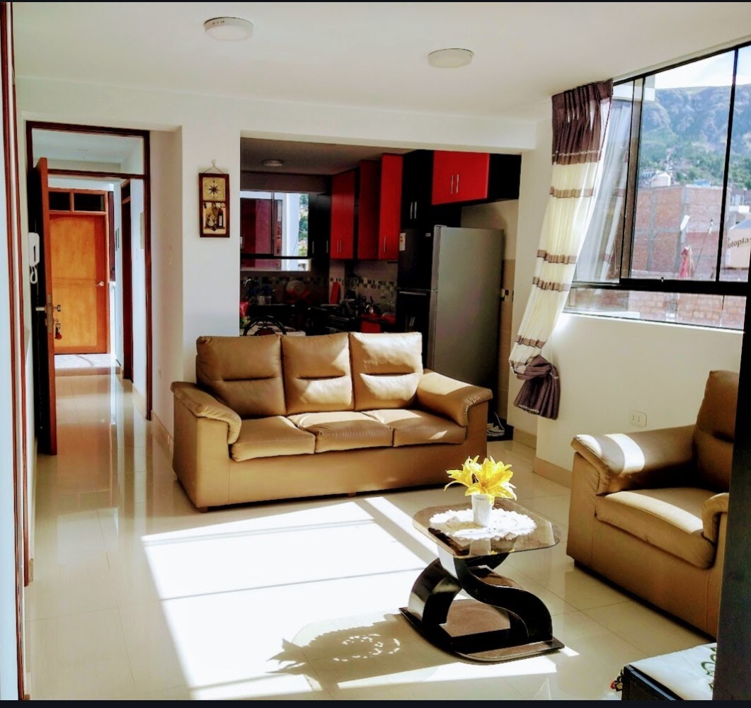 A bright living area features a light-coloured sofa set and a side chair placed around a coffee table. Large windows allow natural light to illuminate the room, while a glimpse of the kitchen area is visible in the background.