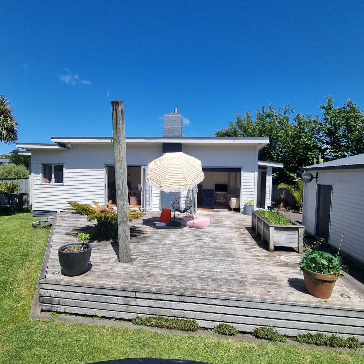 A spacious outdoor deck is featured, with a large umbrella providing shade over colorful lounge seating. Planters filled with greenery and a raised vegetable bed enhance the welcoming atmosphere. The home is set against a clear blue sky, presenting a relaxed outdoor living space.