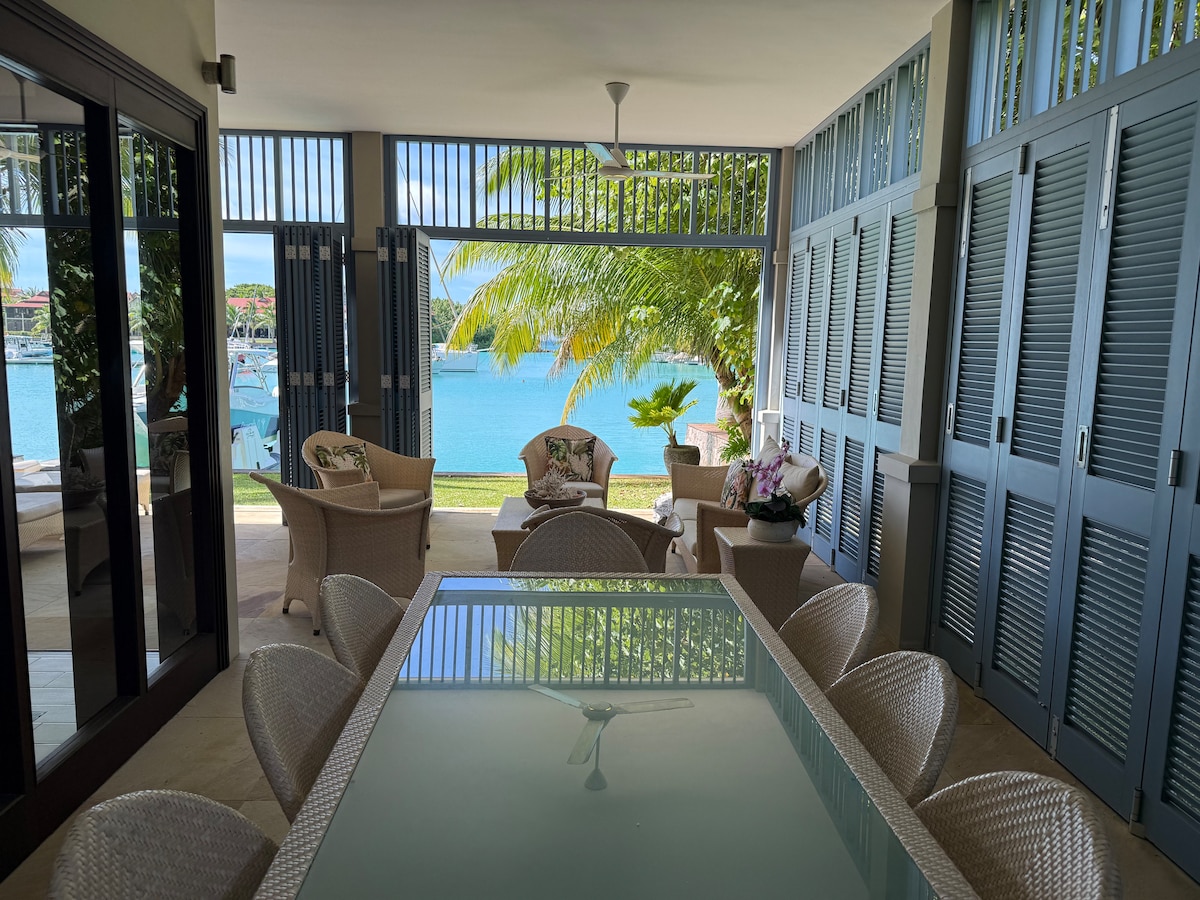A covered terrace features a glass-top dining table surrounded by woven seating. Large windows and louvered shutters frame the stunning view of the marina and sea beyond, complemented by lush palm trees providing a serene ambiance.