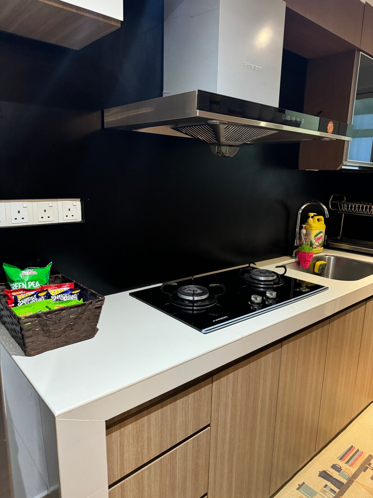 A modern kitchen features a sleek countertop with a built-in gas stove and range hood. A small basket of snacks is set on the surface beside the sink, which reflects a streamlined design with warm wood cabinetry.