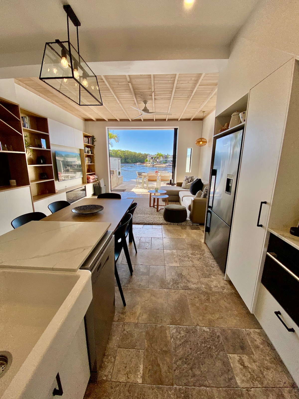 A modern kitchen is visible, featuring a large island with seating and a sink. The space opens up to a living area with comfortable seating and a view of the outdoor deck and water beyond. Natural light floods in through expansive sliding doors.