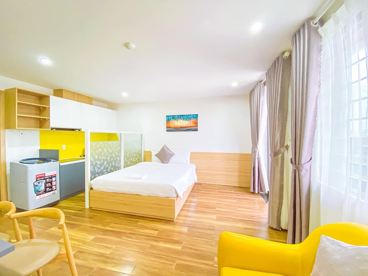 A bright studio apartment features a spacious bed positioned against a wooden wall. Natural light flows through large, sheer curtains. A kitchenette with modern appliances is visible alongside a cozy seating area, complemented by light wood flooring and a vibrant yellow backsplash.