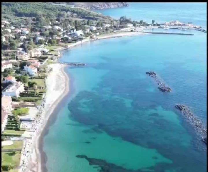 An aerial view captures the serene coastline of Perd'e Sali. The clear waters exhibit various shades of blue and green, while the sandy beach is bordered by lush greenery. The port and nearby structures are visible along the horizon, contributing to the tranquil landscape.