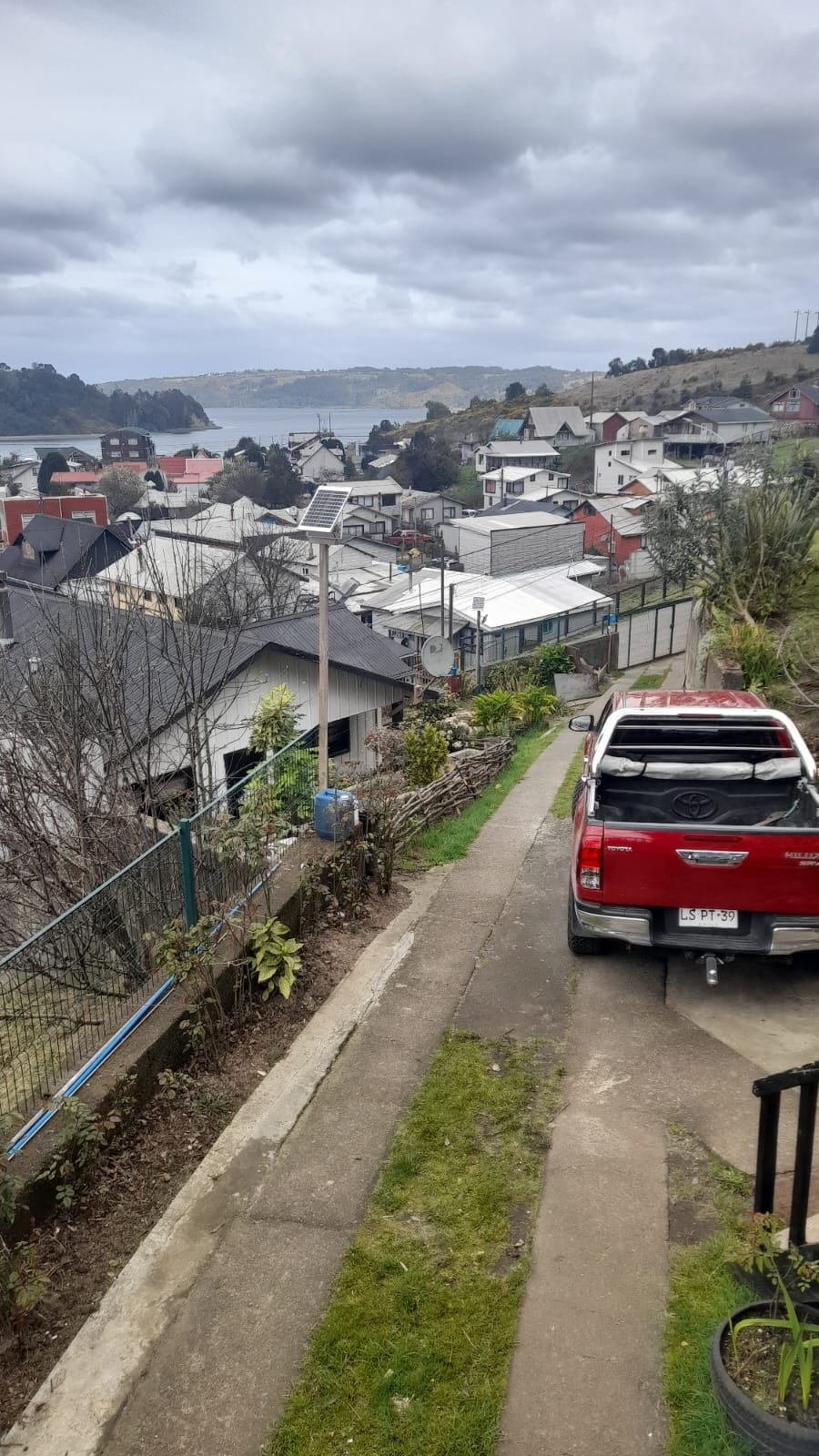 A driveway is seen leading towards a red vehicle parked alongside a well-maintained garden. The view below captures a series of houses with varying rooftops, while the Dalcahue Canal glimmers in the background, framed by lush greenery.