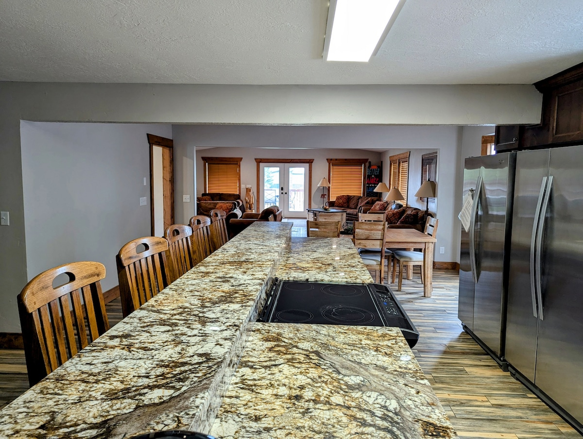 A spacious kitchen is presented with a long, polished granite countertop and multiple wooden barstools along one side. The view extends into a cozy living area with natural light filtering through nearby windows, enhancing the open-concept layout.