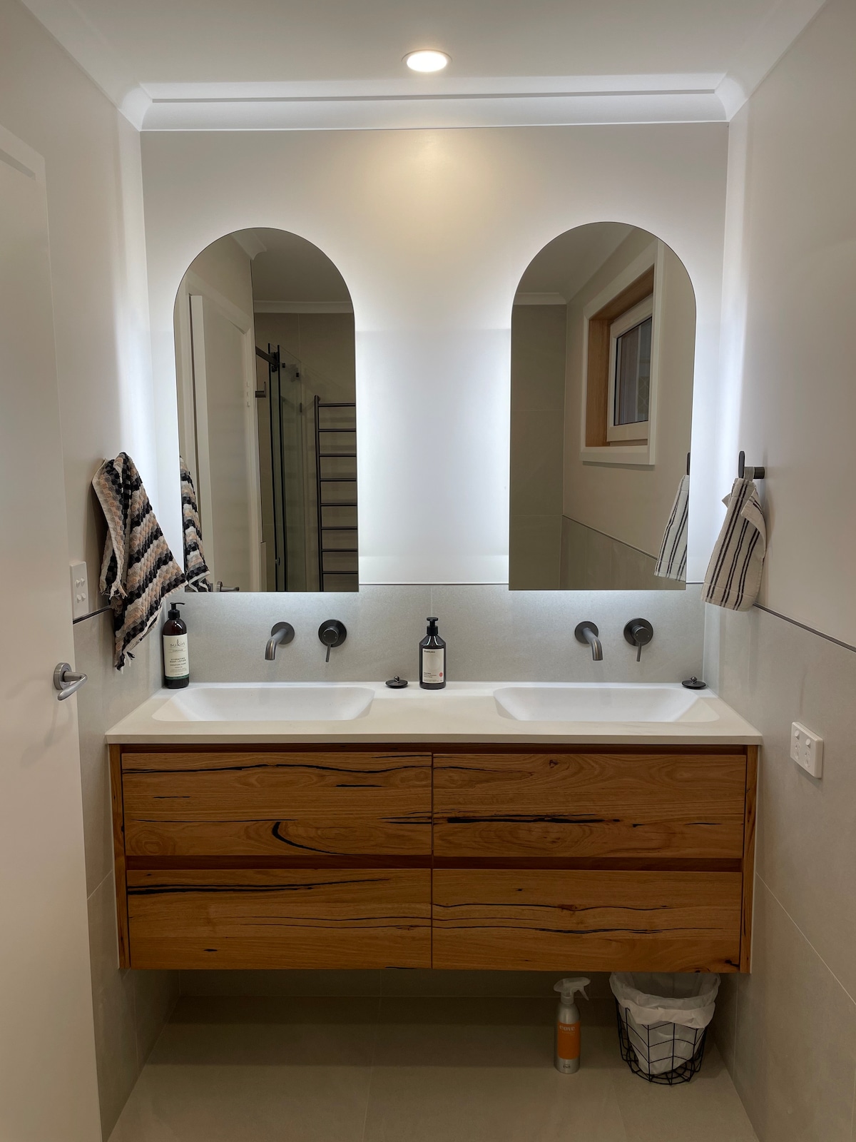 A modern bathroom features a double vanity with wooden cabinetry, framed by two arched mirrors. Wall-mounted faucets and a clean countertop create a minimalist look. Natural light fills the space, complemented by neutral-toned tiles and soft lighting.