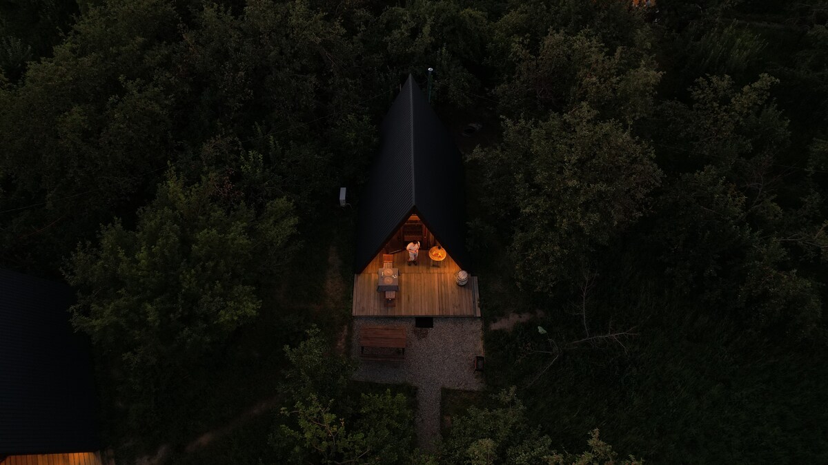 An A-frame cabin is nestled among lush greenery, illuminated softly by warm light. The spacious wooden deck features outdoor seating and a table, while a pathway leads through the surrounding foliage, enhancing the serene and private atmosphere of the location.