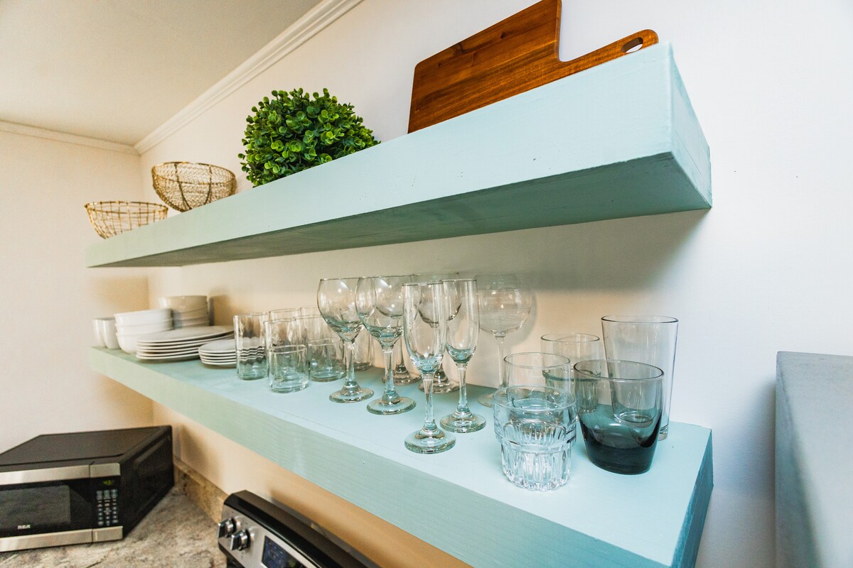 Open shelving displays a collection of glassware, including wine glasses and tumblers, alongside stacked plates. A wooden cutting board and decorative greenery add subtle accents. A microwave is positioned beneath the shelves on a stone countertop.