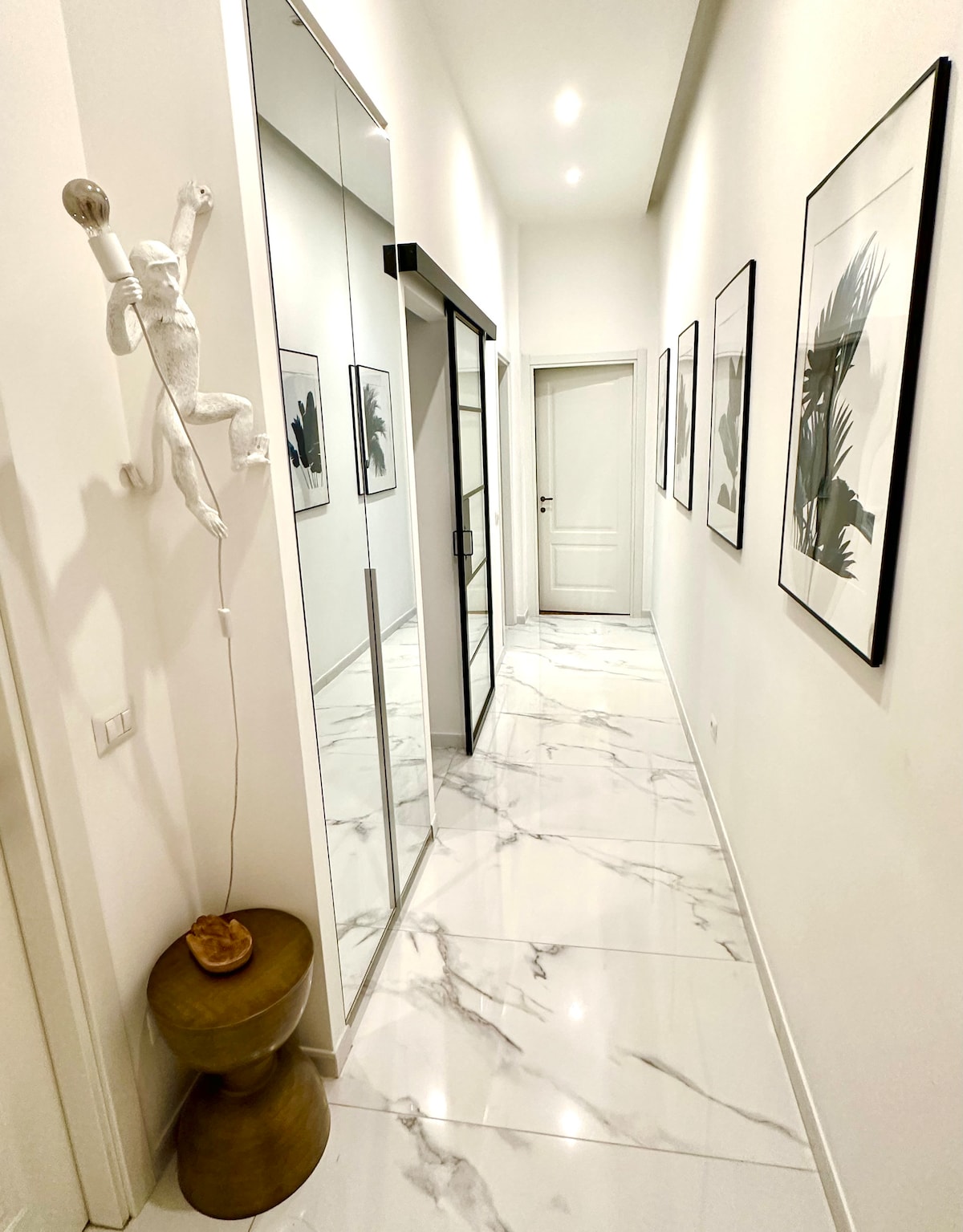 A bright hallway is displayed, featuring a marble floor and white walls adorned with framed botanical prints. A tall mirrored cabinet enhances the sense of space, while a small stool with a decorative object sits beside the wall, adding a touch of design.