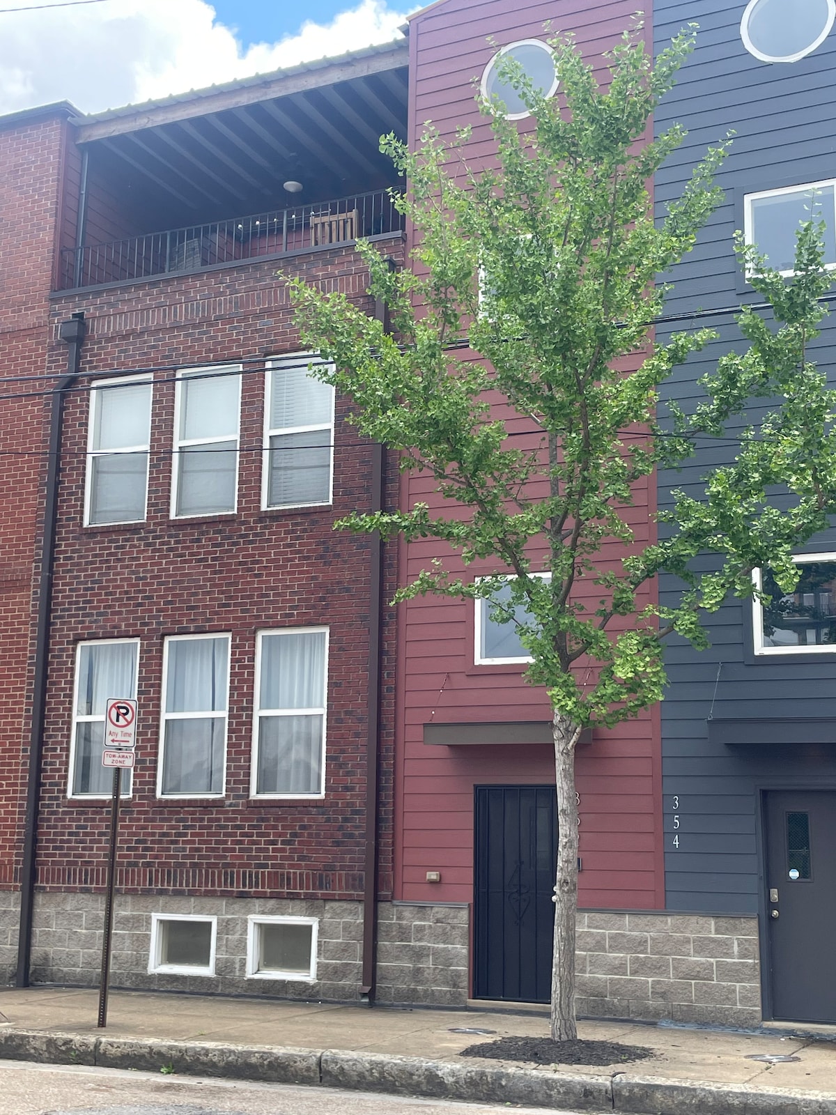The exterior of a four-story townhome is showcased with a mix of red brick and modern siding. Large windows allow natural light, while a tree adds greenery to the entrance. A private patio is visible on the top level, enhancing the urban landscape.