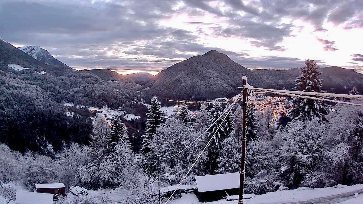 A winter landscape showcases snow-covered mountains and trees, with a serene valley visible below. The scene includes a hint of distant settlements illuminated by soft twilight. Power lines and rustic structures are positioned in the foreground, integrating the natural and built environment.