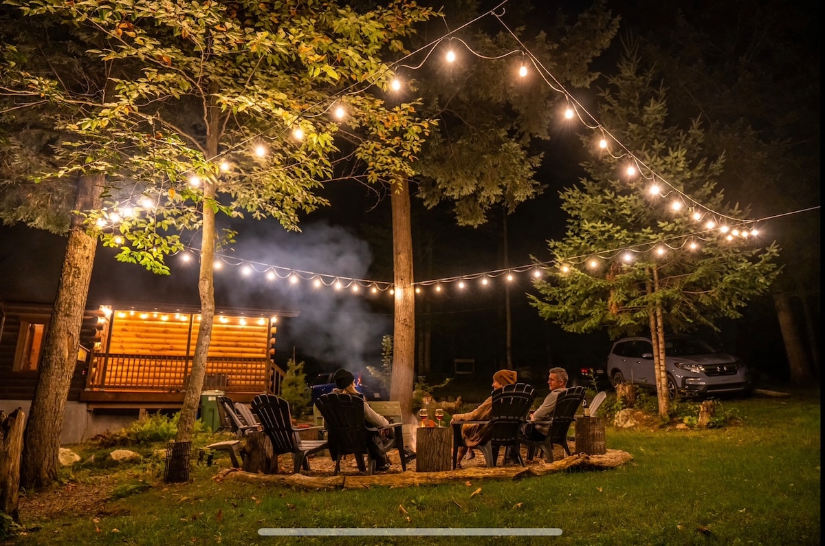 A cozy outdoor seating area is illuminated by warm string lights, creating a welcoming ambiance. Adirondack chairs are arranged around a fire pit surrounded by trees. A cabin is visible in the background, enhancing the peaceful winter retreat atmosphere.