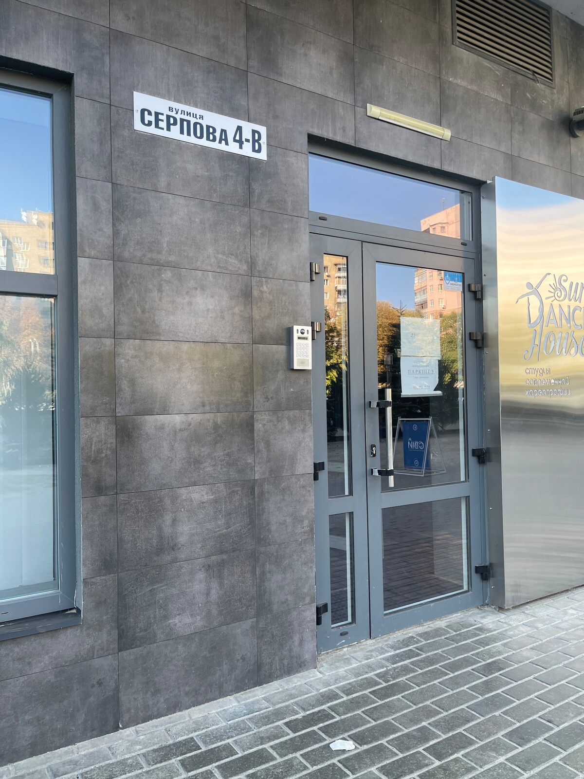 The main entrance to the building is depicted, featuring a gray facade and large glass doors. A sign indicating the address 'Серпова 4-B' is visible above the entrance. The surrounding area is paved with stone, complementing the modern architectural design.