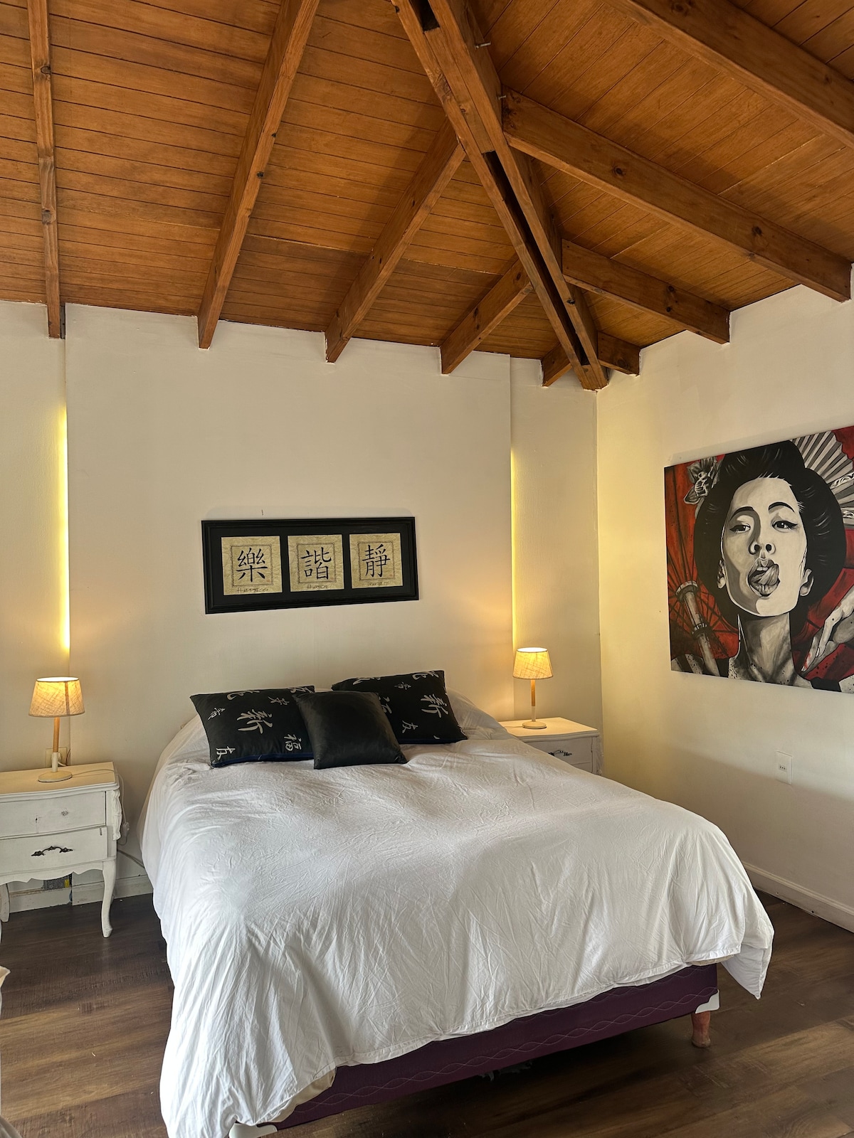 The sleeping area features a spacious bed dressed in crisp white linens and accented with dark pillows. Two bedside tables with lamps stand on either side. Natural wood beams create an airy ceiling, and a piece of artwork adorns the wall, adding a touch of character.