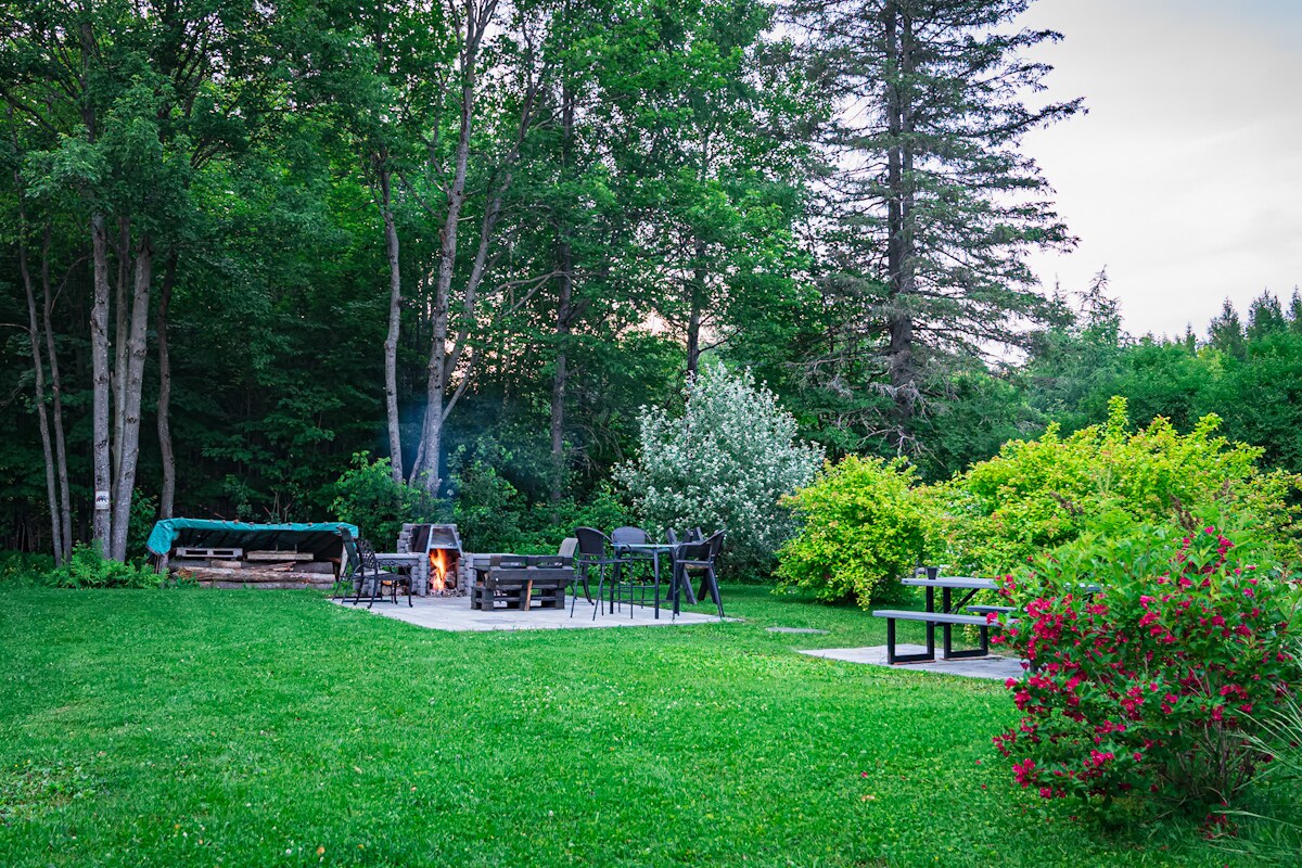 An outdoor area features a fire pit surrounded by chairs, set on a grassy expanse. A wooden seating area is positioned nearby, with stack of firewood and lush greenery, including trees and flowering shrubs, providing a peaceful natural backdrop.