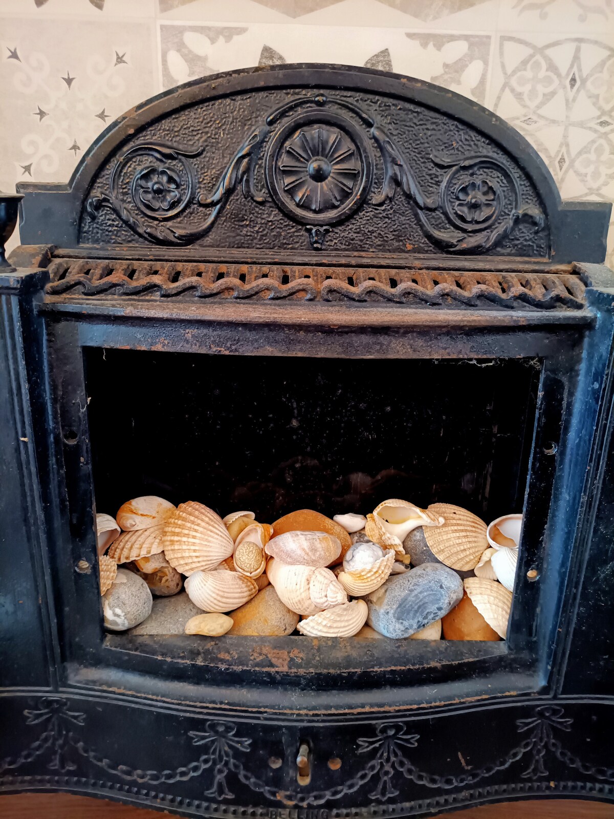 An antique fireplace is showcased, its ornate design highlighted by intricate detailing. Inside, a collection of seashells and smooth stones creates an organic display, blending natural elements with the historic structure.