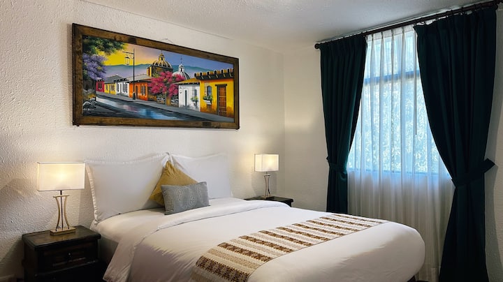Een lichte en elegante kamer met een tweepersoonsbed, decoratie geïnspireerd door La Antigua Guatemala en een schilderij van een straat in de koloniale stad. Het beschikt over een kast en verduisterende gordijnen voor een goede nachtrust.