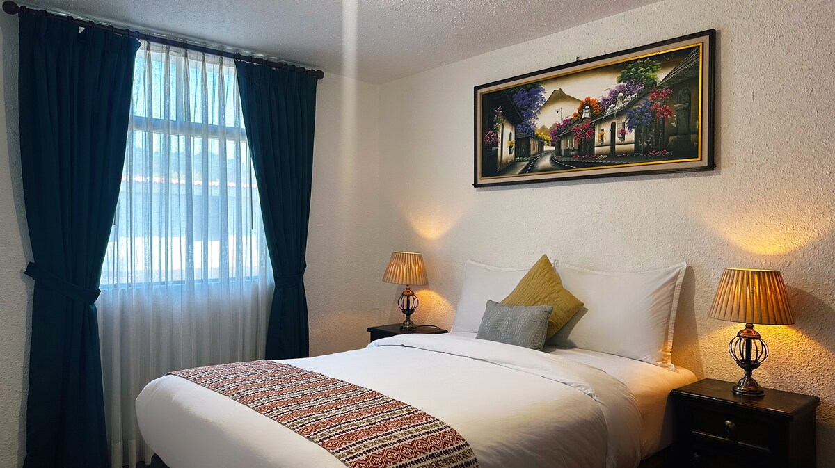 חדר מואר ואלגנטי עם מיטה זוגית, עיצוב בהשראת לה אנטיגואה גואטמלה וציור של הר הגעש כגיבור. הוא כולל ארון ווילונות האפלה לשינה נינוחה.