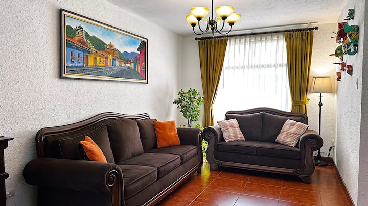 Gezellige woonkamer met een koffiekleurige bank, elegante gordijnen en een muur met kleurrijke dierenmaskers. Een kleine boom voegt een natuurlijke en charmante toets toe aan de sfeer.