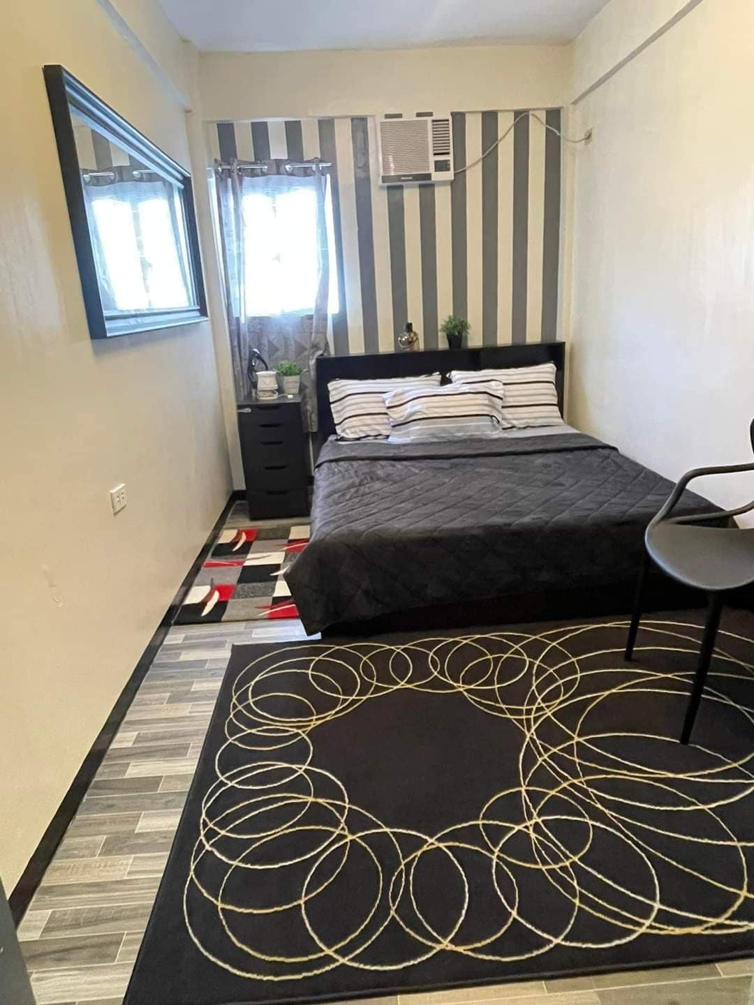 A compact bedroom features a black and white striped accent wall. A spacious bed with striped bedding is positioned against the wall. A small black nightstand and a chair are present, while a geometric rug adds texture to the floor. Natural light enters through a window dressed with light curtains.