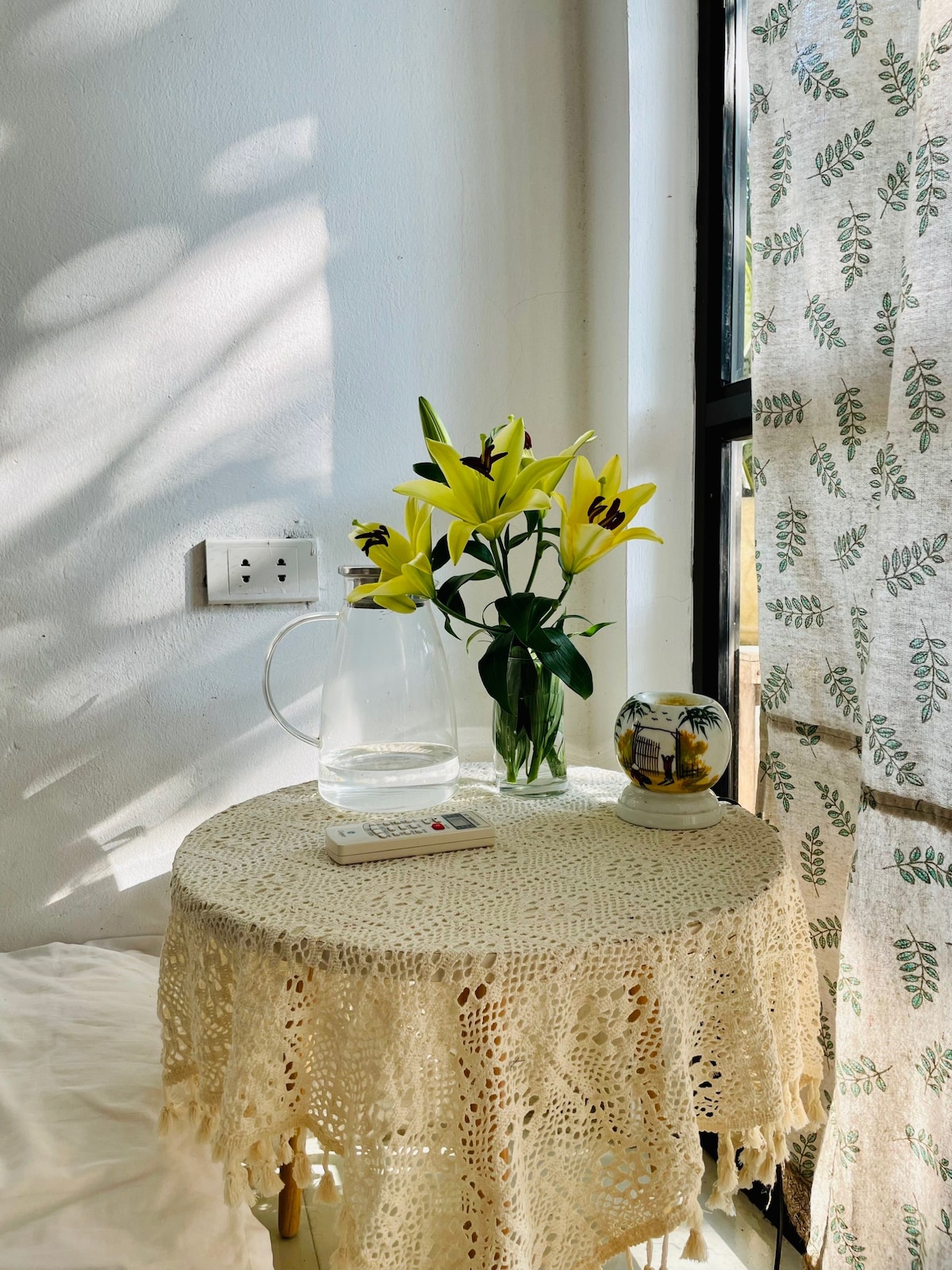 A small, round table is covered by a delicate lace tablecloth, topped with a pitcher of water and a vase of yellow lilies. A decorative orb sits next to the vase. Sunlight filters through a semi-transparent curtain adorned with leafy patterns, creating soft shadows.