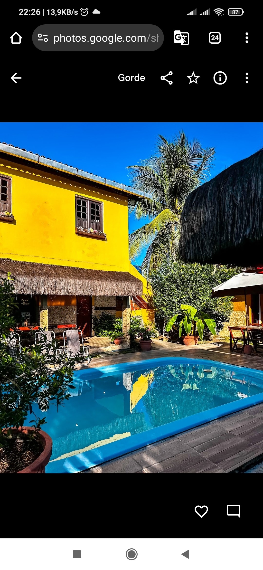 A vibrant yellow structure captures attention in the background, surrounded by lush greenery. A sparkling pool reflects the clear blue sky, with lounge chairs placed nearby. A shaded outdoor seating area is visible, offering a comfortable space to relax by the pool.
