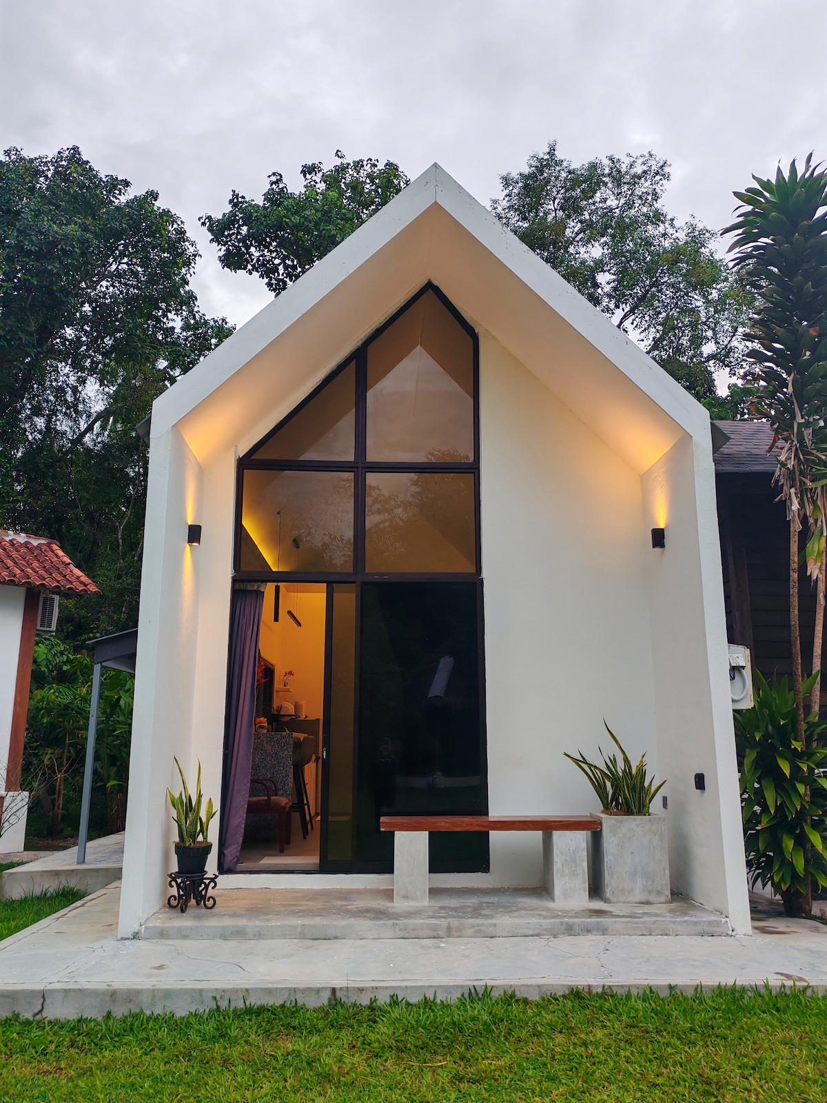 The exterior of a modern chalet features a triangular roof and large glass windows that allow natural light to fill the interior. Surrounded by greenery, a small bench sits on a concrete porch, enhancing the connection with the peaceful outdoor setting.
