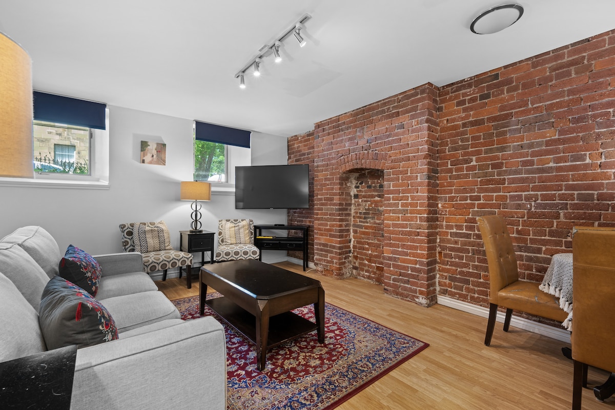 Exposed brick walls create a warm backdrop for the cozy living area, which includes a light-colored sofa and two patterned chairs. A dark wood coffee table centers the space, accompanied by a wall-mounted television, while a dining area with two chairs is visible in the corner.