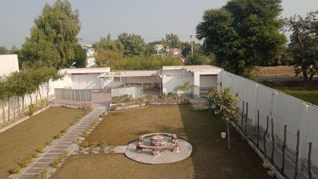 A well-maintained outdoor area is showcased, featuring a circular stone seating area surrounded by greenery. Neat pathways made of stones lead across the manicured lawn, with trees lining the property. White walls enclose the space, providing a sense of privacy.