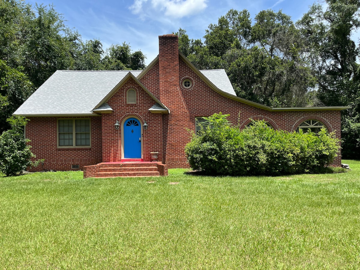 A charming brick house is presented, featuring a distinctive blue front door. Lush greenery surrounds the home, with well-maintained shrubs and a spacious front yard of vibrant grass. Large windows with rounded details add character to the structure, complemented by a traditional chimney.