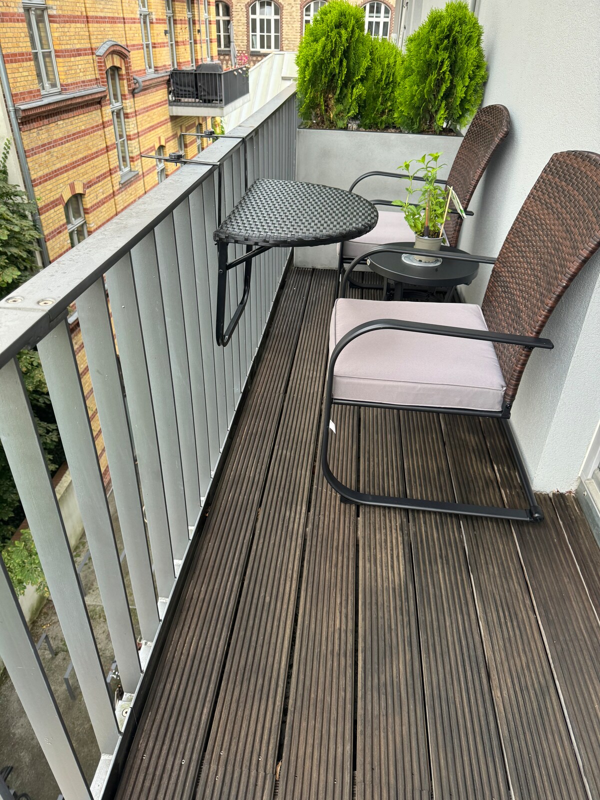 A balcony space is presented with a small round table and two chairs, complemented by a potted plant on the table. The wooden decking offers a warm ambiance, while the surrounding greenery adds a touch of nature to the urban setting.