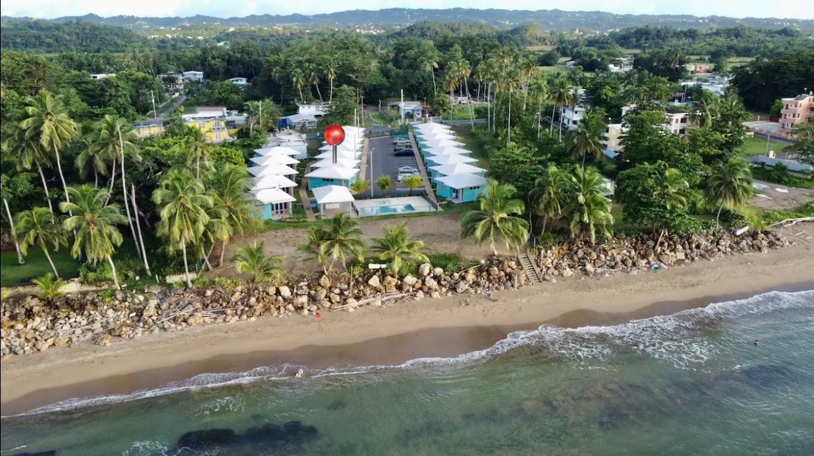 Aerial view captures the property nestled along the beach, with well-maintained bungalows surrounded by palm trees. A shared swimming pool is visible in the center, complementing the oceanfront setting and sandy shoreline that merge with gentle waves.