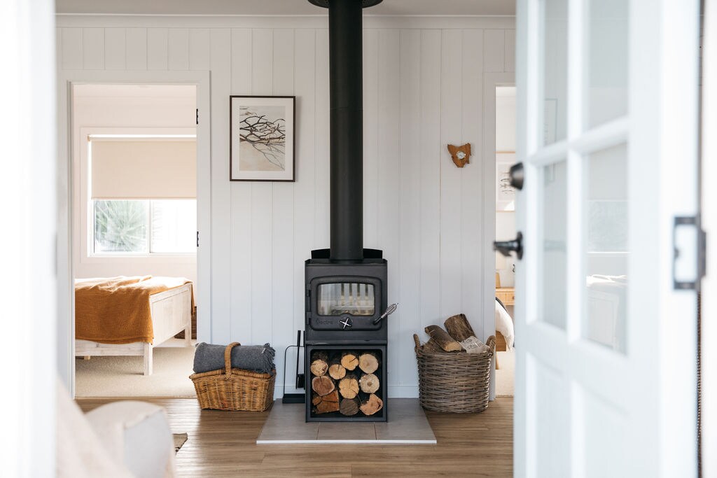A wood-burning stove is positioned at the center of the open space, surrounded by neatly stacked firewood. A woven basket holds extra blankets, while light streams through windows, illuminating the white walls and light wooden flooring.