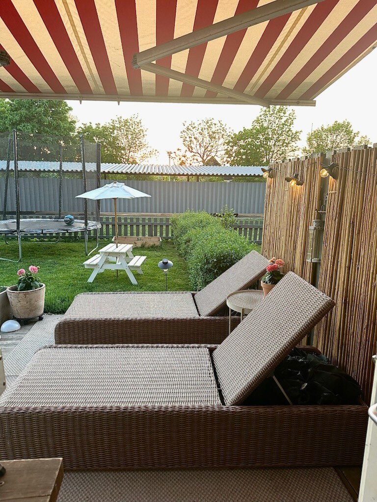 Two lounge chairs are positioned under a red and white striped awning, facing a grassy area with a picnic table. Soft lighting from nearby fixtures creates a relaxing ambiance as the sun sets in the background.
