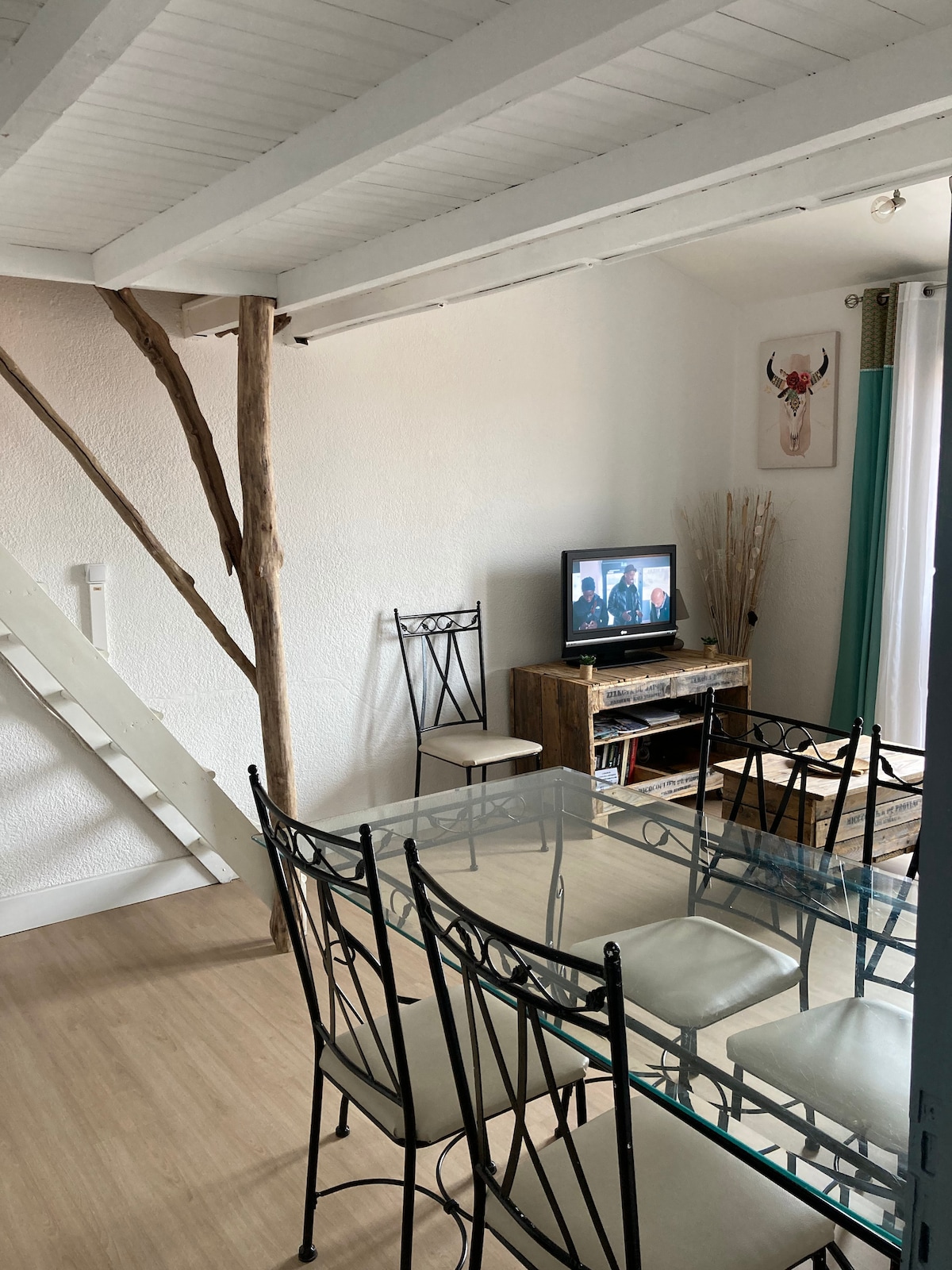 The living area features a glass dining table surrounded by six metal chairs, with a wooden media console displaying a television. A staircase leads to the mezzanine, and natural light enters through a window adorned with green curtains, enhancing the room's brightness.