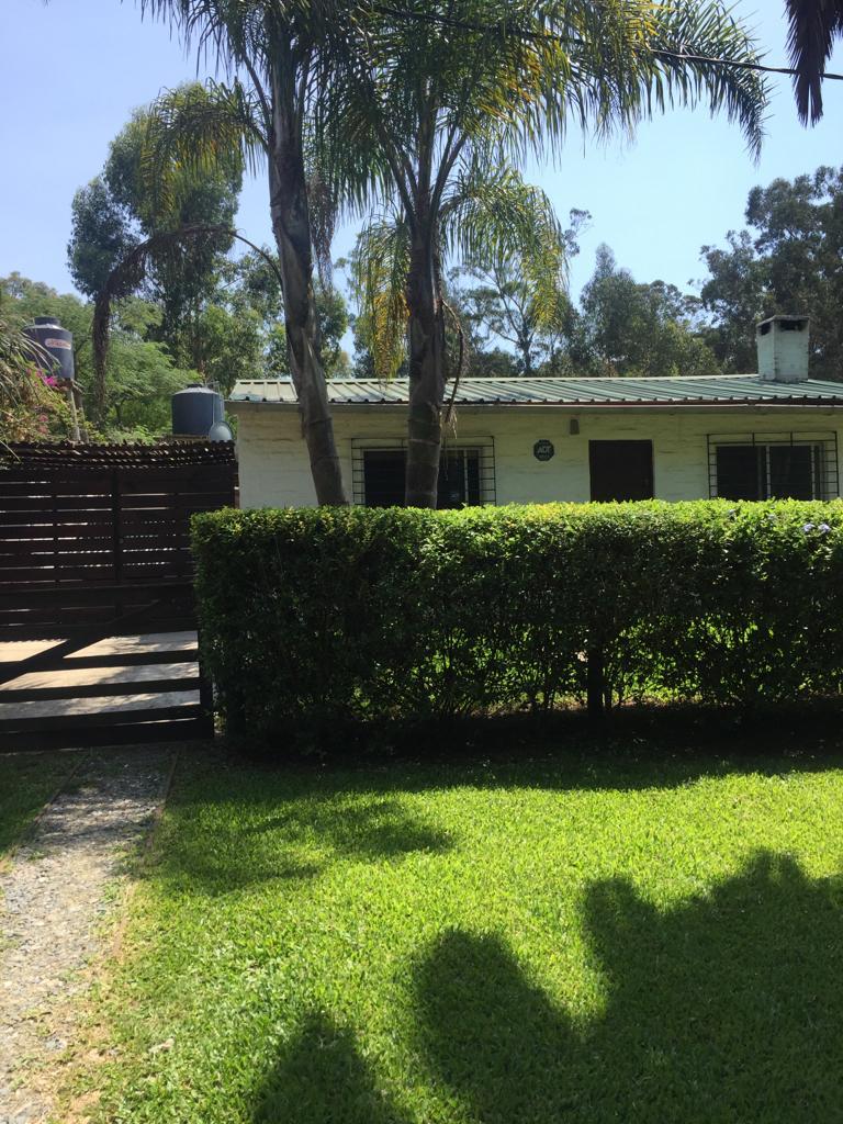 A single-story home is visible, surrounded by greenery and manicured grass. A wooden fence is present in the foreground, offering privacy. Two palm trees can be seen flanking the house, which features large windows and a sloped roof.