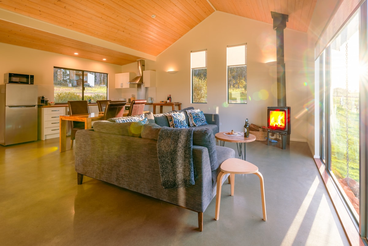 A spacious living area showcases a cozy lounge with a plush sofa and a wooden coffee table. A Norwegian wood-heater is positioned near large windows that allow natural light to fill the room, providing views of the surrounding landscape, while a modern kitchen is visible in the background.