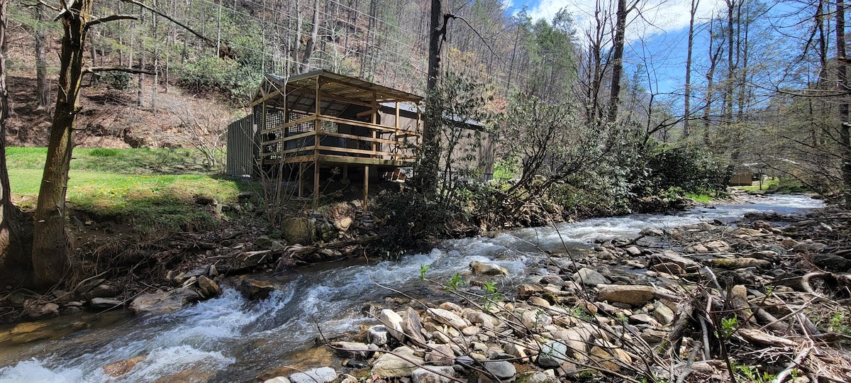 A tiny cabin overlooks a gently flowing stream, surrounded by natural terrain. The cabin features a spacious deck, offering views of the water and nearby trees. Sunlight breaks through the canopy, highlighting the lush greenery along the stream’s bank.