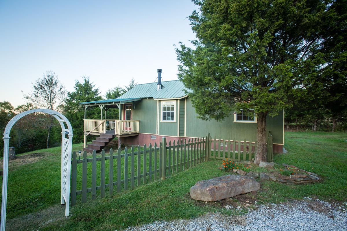 The cabin's exterior is framed by a green facade and a sloped metal roof. A welcoming porch with steps leads to the entrance, flanked by a manicured lawn and mature trees. A white archway stands at the entrance of a gravel driveway, enhancing the inviting landscape.