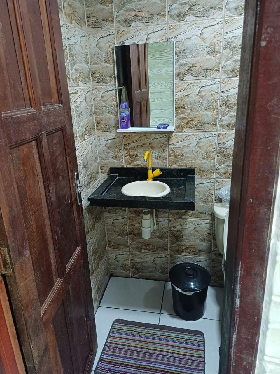 A compact bathroom is shown featuring a dark granite countertop with a circular sink. A mirror is mounted above the sink, and a small black wastebasket is positioned nearby. The walls are tiled with a stone pattern, and a colorful area rug is placed on the floor.