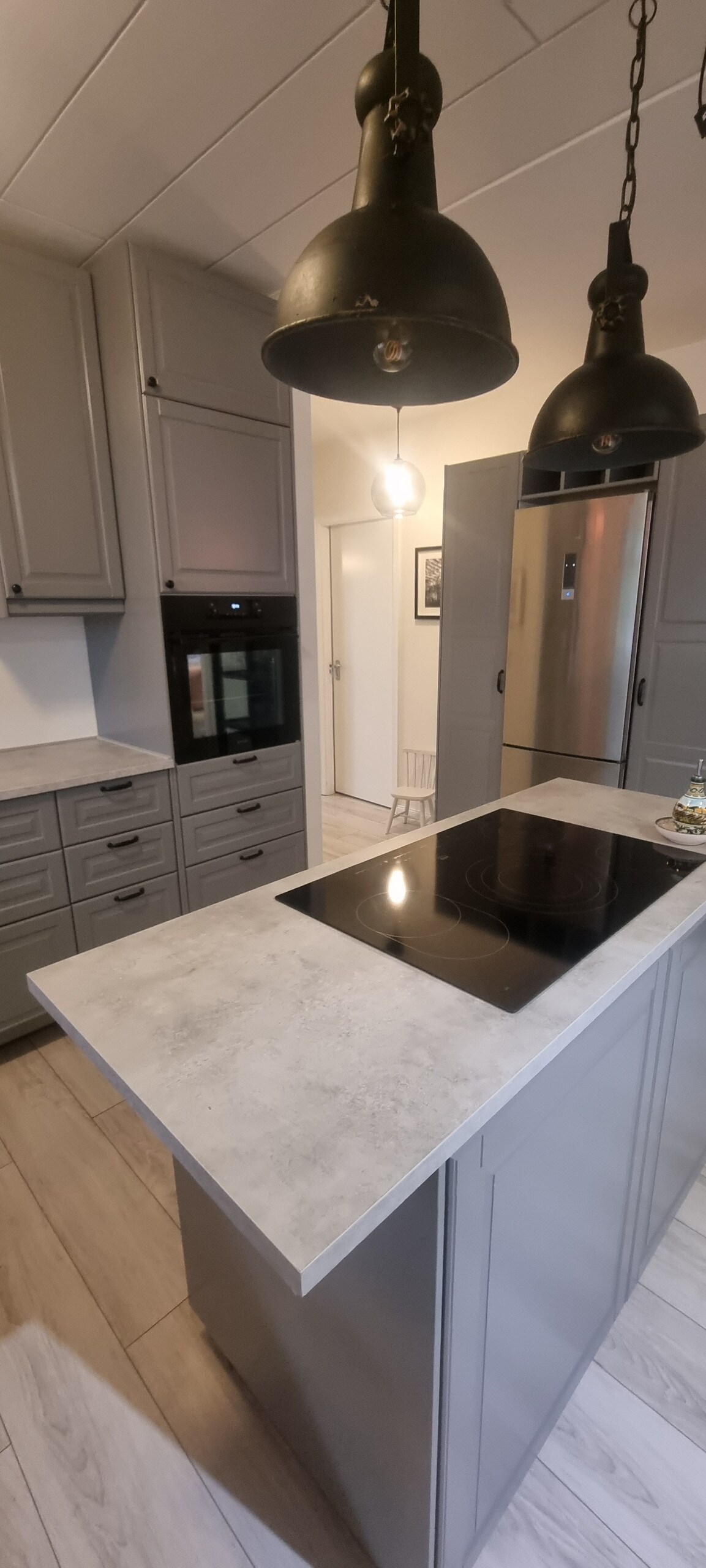 A modern kitchen features sleek gray cabinetry and a light-colored countertop. An integrated oven and refrigerator are visible, along with a black cooktop on the island. Pendant lights hang above the kitchen island, providing illumination. The space is complemented by plank flooring.
