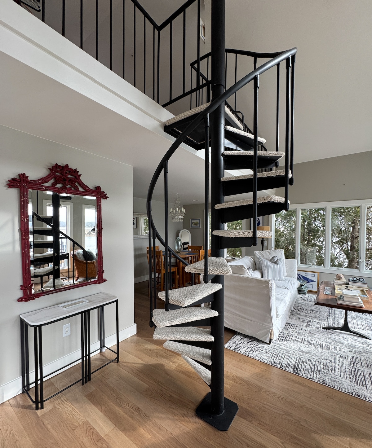 A spiral staircase with a sleek black railing leads to the loft bedroom. A mirror with a burgundy frame is mounted against the wall, reflecting the light. The open living area features a light-colored couch and a wooden dining table, visible through large windows offering views of the outdoors.