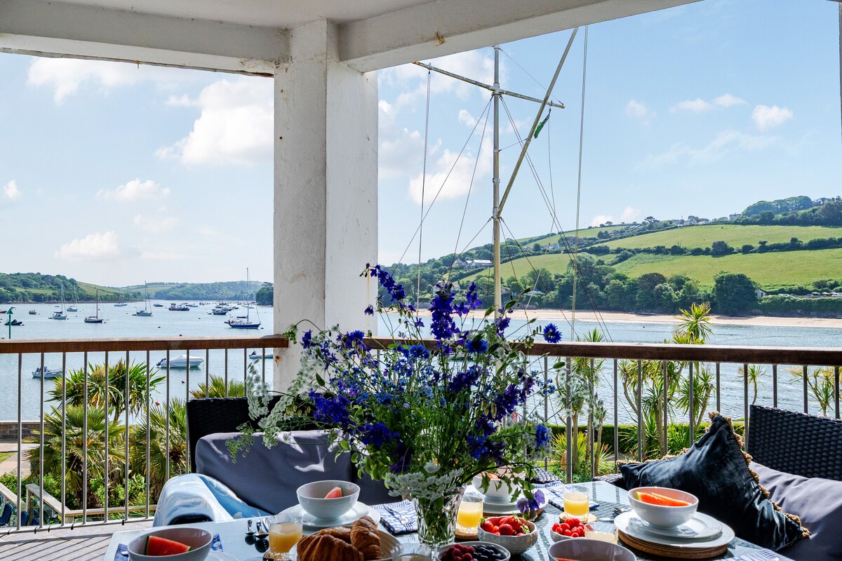 A panoramic balcony is featured, overlooking the scenic Salcombe estuary. A dining table is set with bowls of fruit, pastries, and beverages. Vibrant blue flowers are arranged in the center, complementing the tranquil view of boats and green hills in the background.