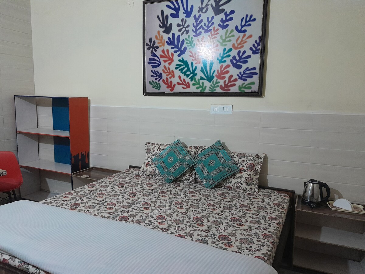 A spacious bedroom features a patterned bedspread and two decorative cushions against a backdrop of colorful wall art. A small shelf and a bedside table are visible, with a kettle and cup positioned neatly. A chair adds functional seating to the room.