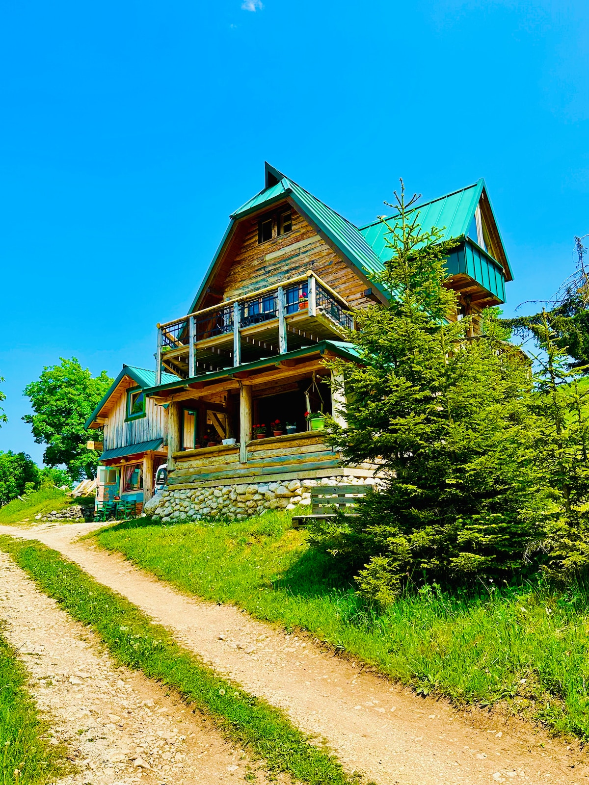 A charming wooden cabin is set along a gravel path, surrounded by greenery. The structure features a sloping green roof and a spacious balcony adorned with railings. Nearby coniferous trees add to the natural setting, contributing to the serene atmosphere of the area.