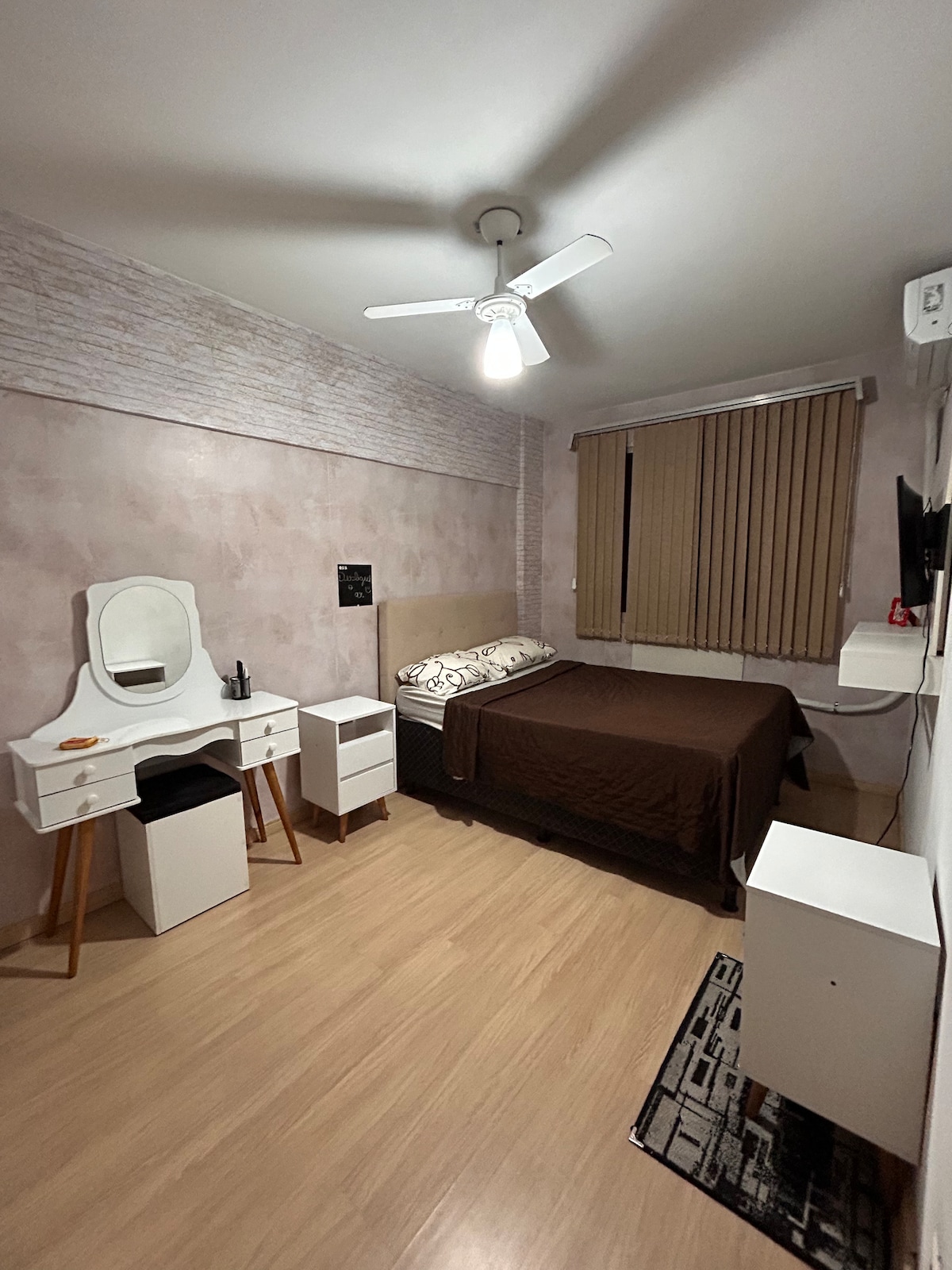 A cozy bedroom is presented with a spacious bed covered in a brown blanket. A ceiling fan provides airflow, while a window with vertical blinds allows for light control. A small vanity and a white nightstand are included, contributing to the room's functionality.