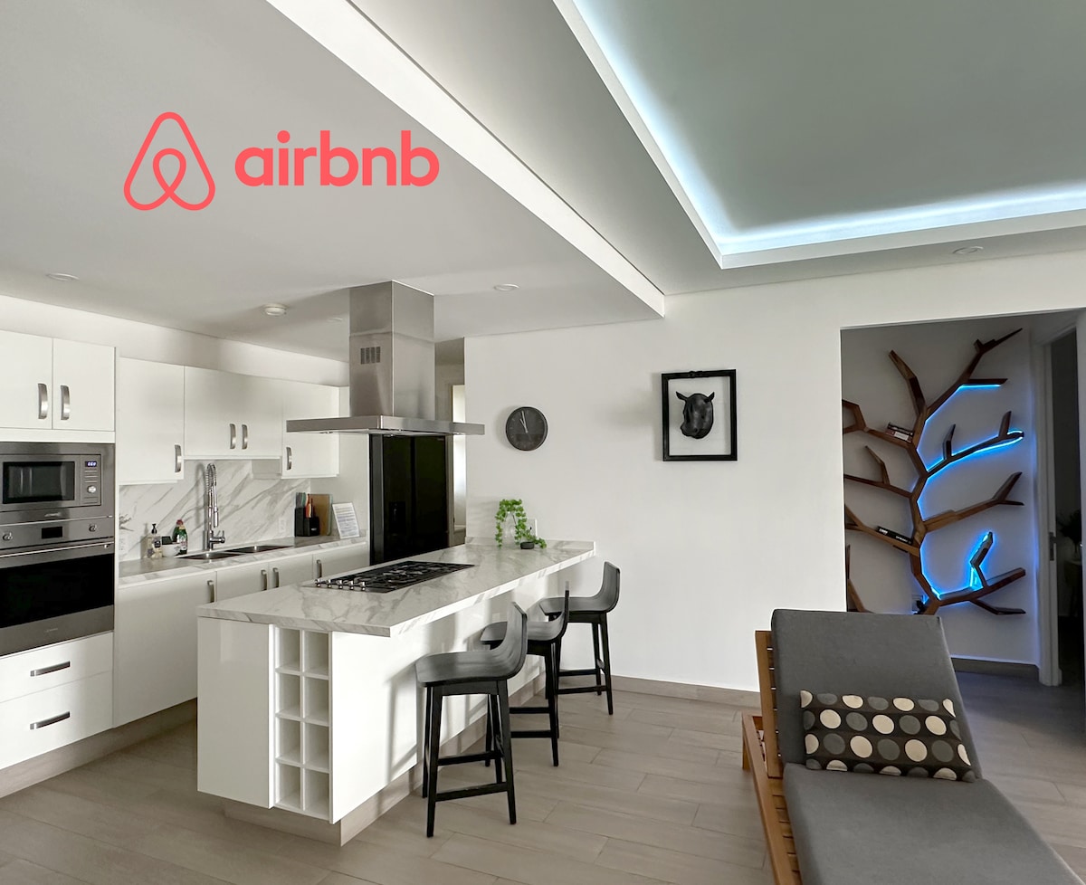A modern kitchen area features sleek white cabinetry and a marble countertop. Barstools are positioned at the counter, with a built-in stove and microwave visible. A unique wall sculpture with integrated lighting adds an artistic touch to the space.