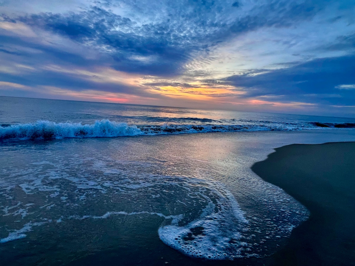 The serene seascape captures gentle waves lapping at the shore, with soft, foamy edges reflecting the colors of a fading sunset. A tranquil sky features a gradient from warm orange to cool blue, creating a peaceful atmosphere over the expansive ocean.