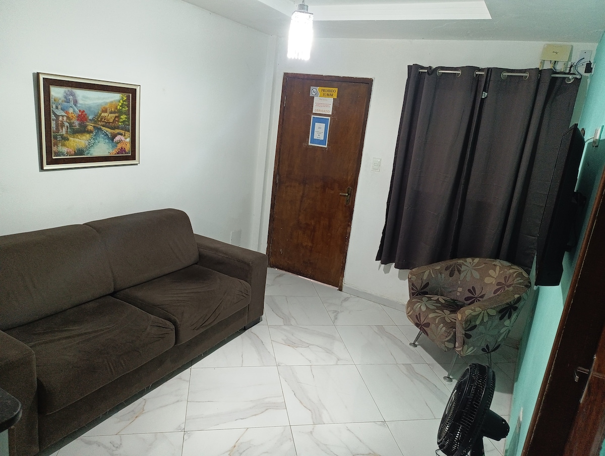 A living area features a brown sofa and a patterned chair, providing seating options. A fan is positioned nearby, and a small side table is visible. Light-colored tiles cover the floor, while dark curtains frame a window. A framed artwork enhances the ambiance.